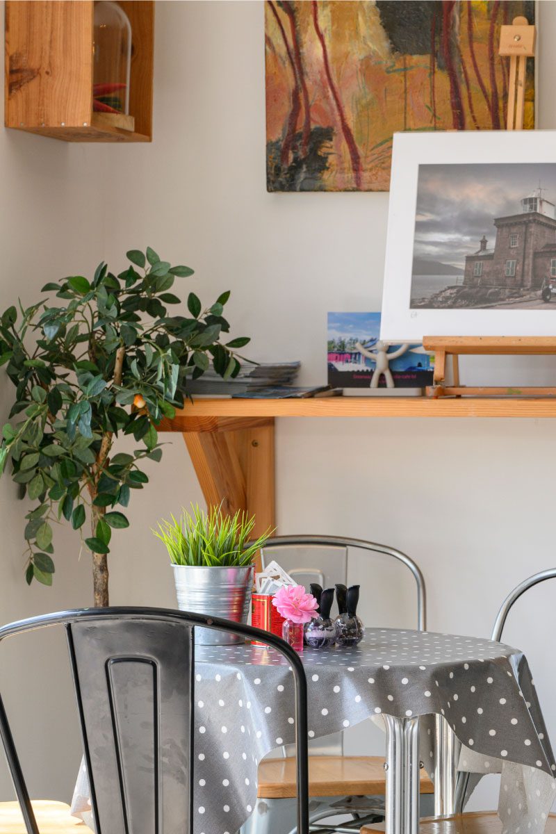 A small café table with a gray polka-dot tablecloth, potted plants, condiments, and napkins. Behind it, a shelf displays artwork from Greenway Lane Art Studio, a potted plant, and stacked plates against a light-colored wall.