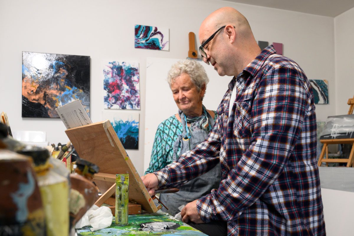 A man and an older woman paint together at an easel in Greenway Lane Art Studio, surrounded by colorful abstract artwork. The man wears glasses and a plaid shirt; the woman, in a patterned top and apron, smiles as they work.