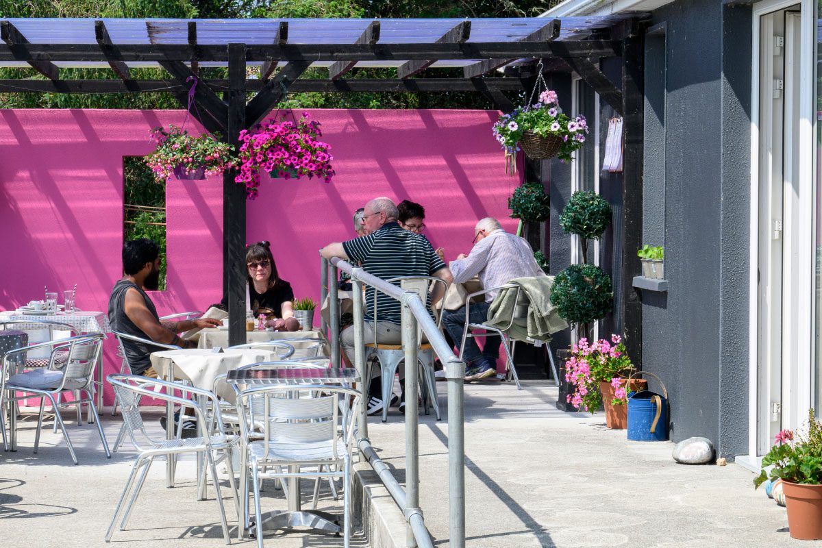 People are dining at outdoor tables on a sunny patio with a bright pink wall, potted plants, and hanging flowers near Greenway Lane Art Studio. Some are eating and talking while others relax quietly in the vibrant atmosphere.