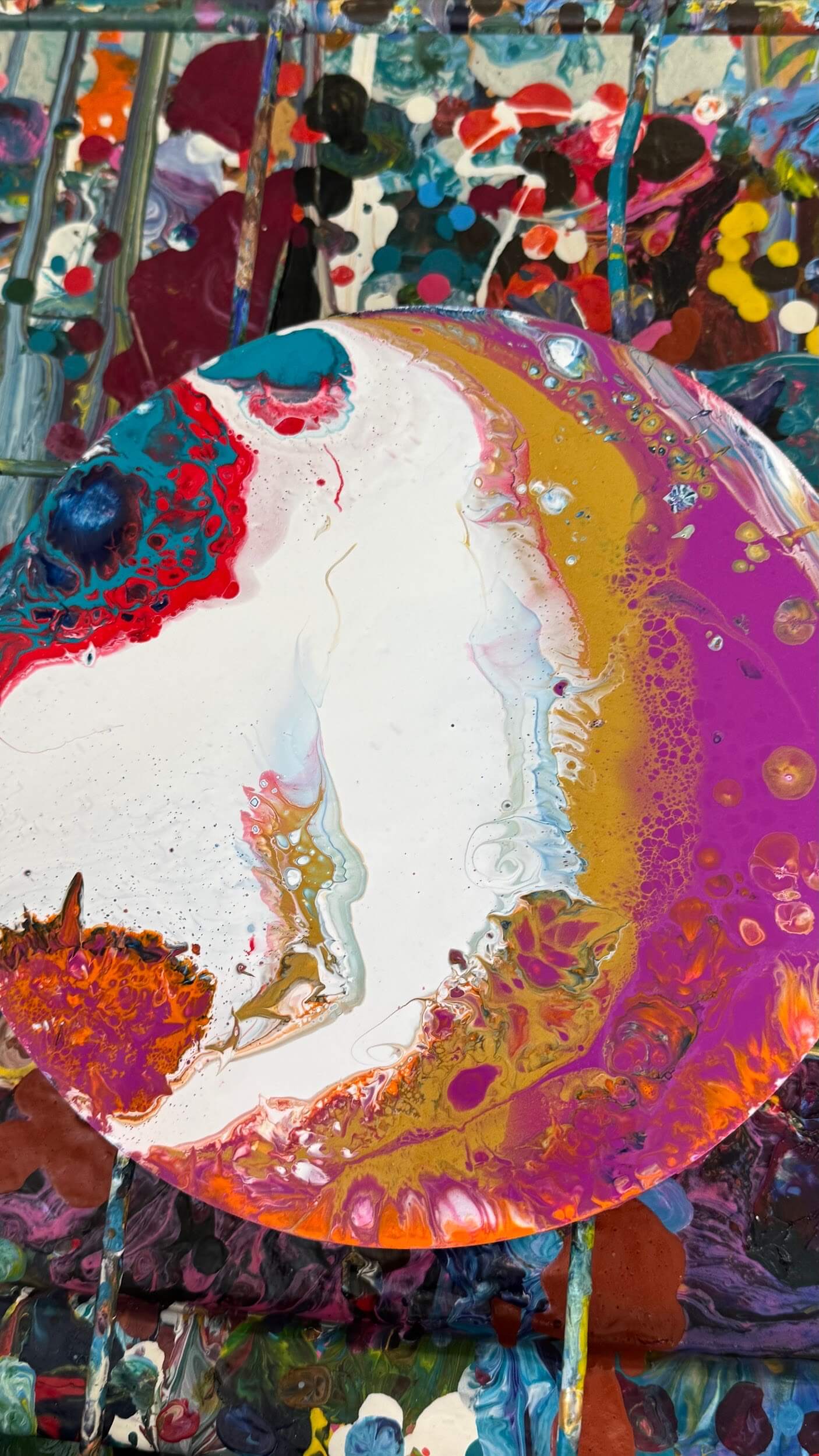 A circular canvas covered in abstract, colorful paint swirls of white, gold, pink, red, and orange rests on a messy, paint-splattered surface at Greenway Lane Art Studio, surrounded by vibrant splashes and blobs of color.