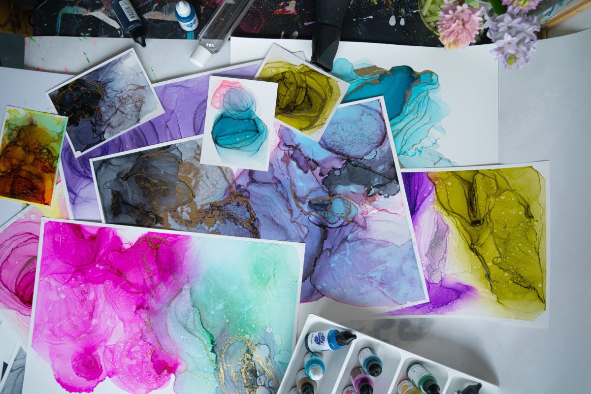 A collection of colorful abstract watercolor paintings, mainly in pink, purple, blue, and yellow hues, is scattered on a table at Greenway Lane Art Studio with paint bottles, art supplies, and flowers in the background.