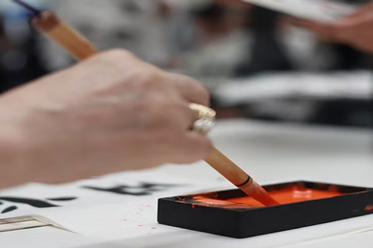 At Greenway Lane Art Studio, a person dips a brush into red ink while practicing calligraphy, with black characters visible on white paper and blurred background elements.