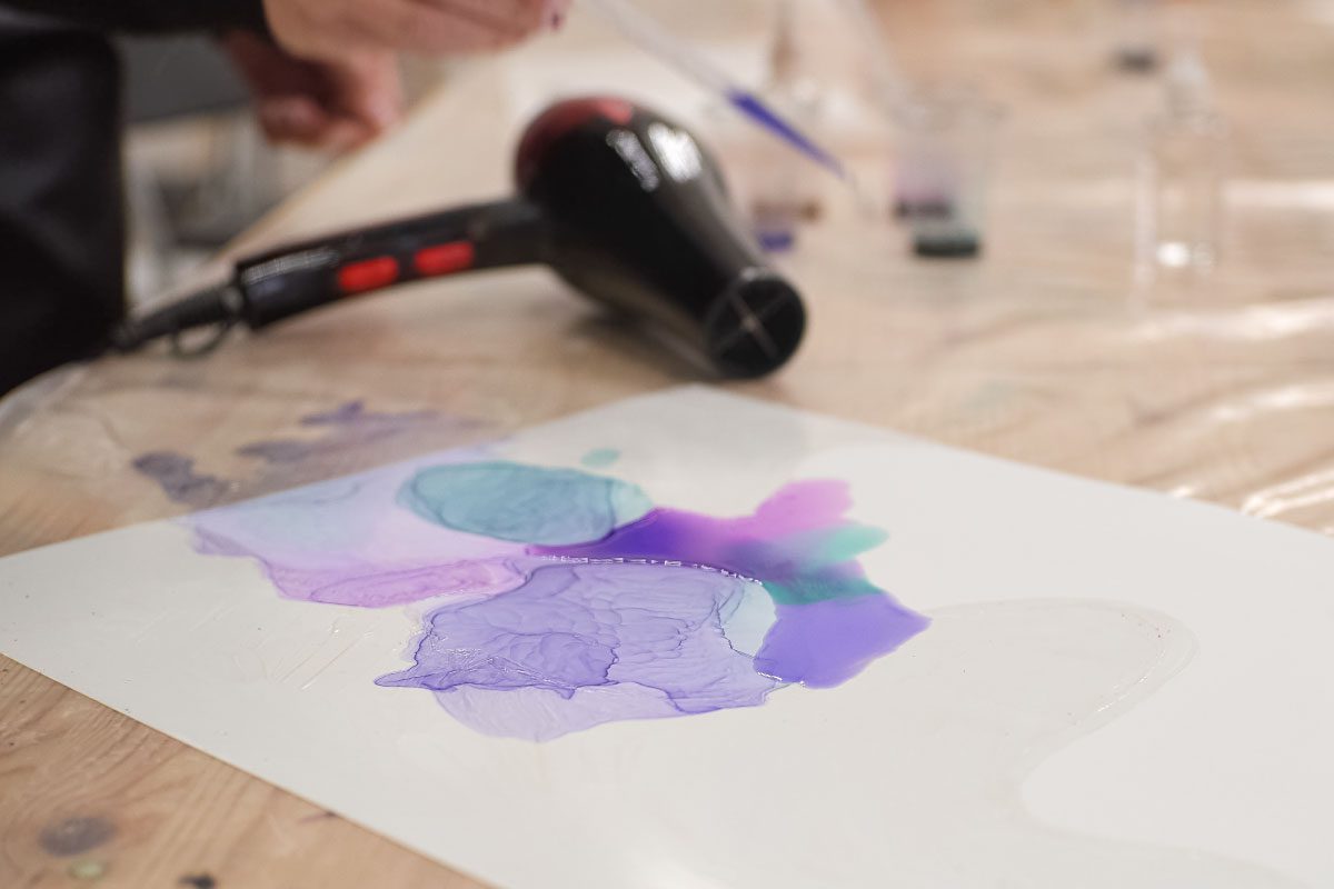 At Greenway Lane Art Studio, a hand uses a pipette to apply liquid on a white sheet with purple and blue stains, while a black hairdryer rests nearby on a wooden table covered with plastic.