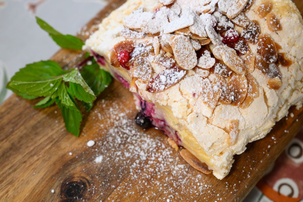 A slice of berry and almond meringue roulade sits on a wooden board at Greenway Lane Art Studio, dusted with powdered sugar and garnished with fresh mint leaves.