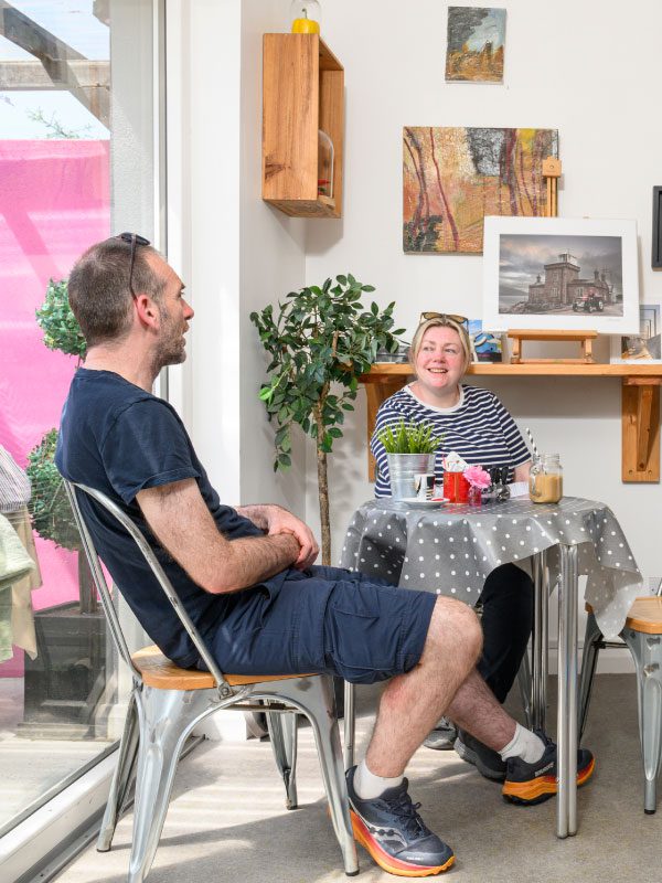 Two people sit at a small table in a bright cafe, smiling and talking. The table is covered with a grey polka dot cloth, flowers, and drinks. Art from Greenway Lane Art Studio and lush plants decorate the walls around them.