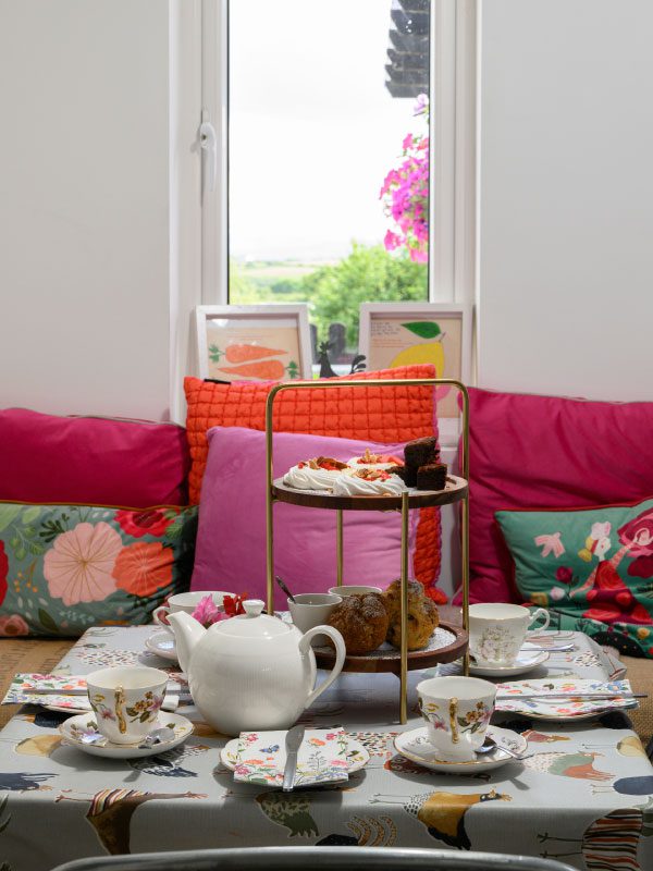 A colorful afternoon tea setup from Greenway Lane Art Studio features floral teacups, a white teapot, and a tiered tray of pastries on a patterned tablecloth. Bright pillows and a window with greenery and flowers complete the scene.