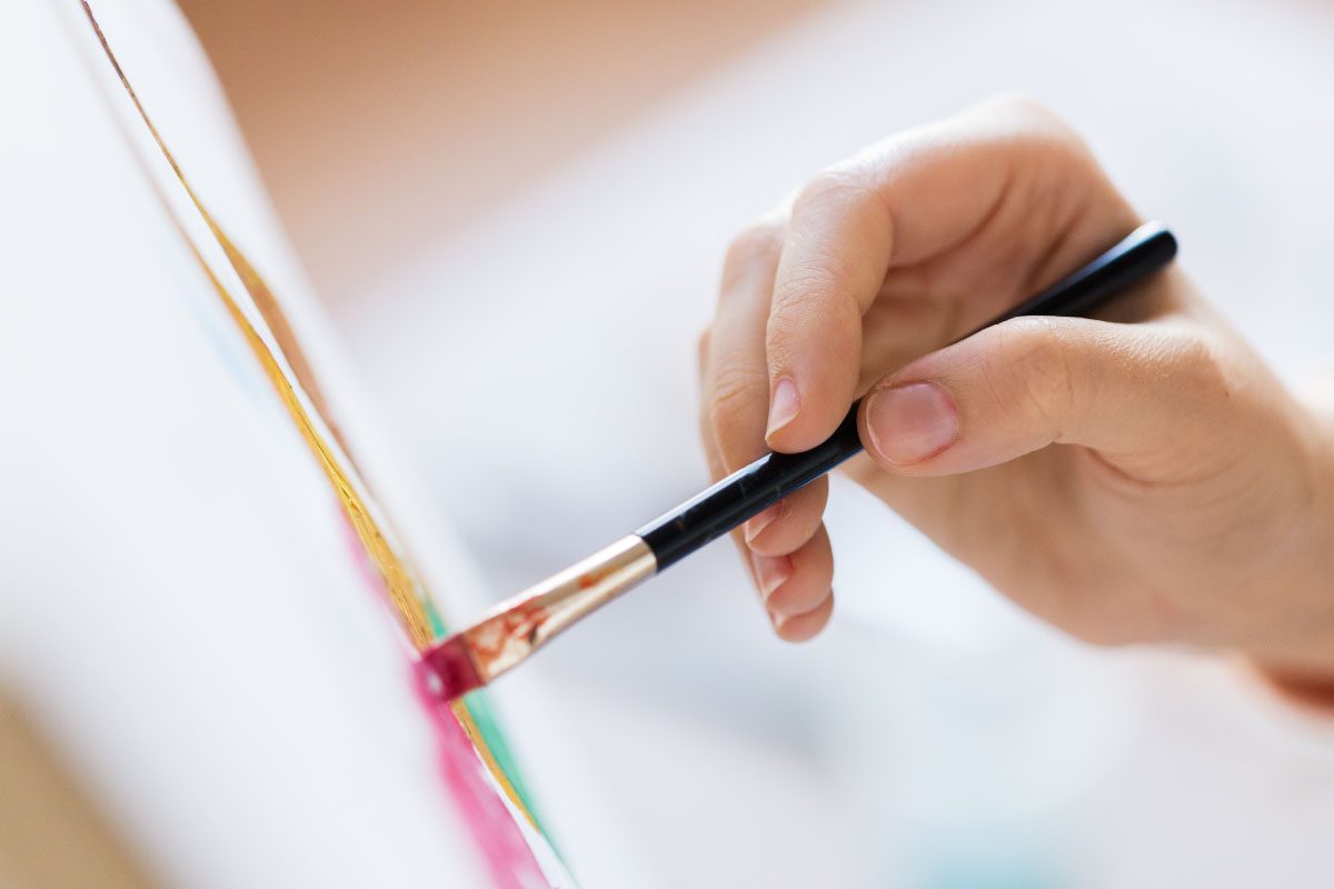 A close-up of a hand holding a paintbrush, applying pink paint to a canvas with gentle, precise strokes. The background is blurred, focusing attention on the painting activity at Greenway Lane Art Studio.