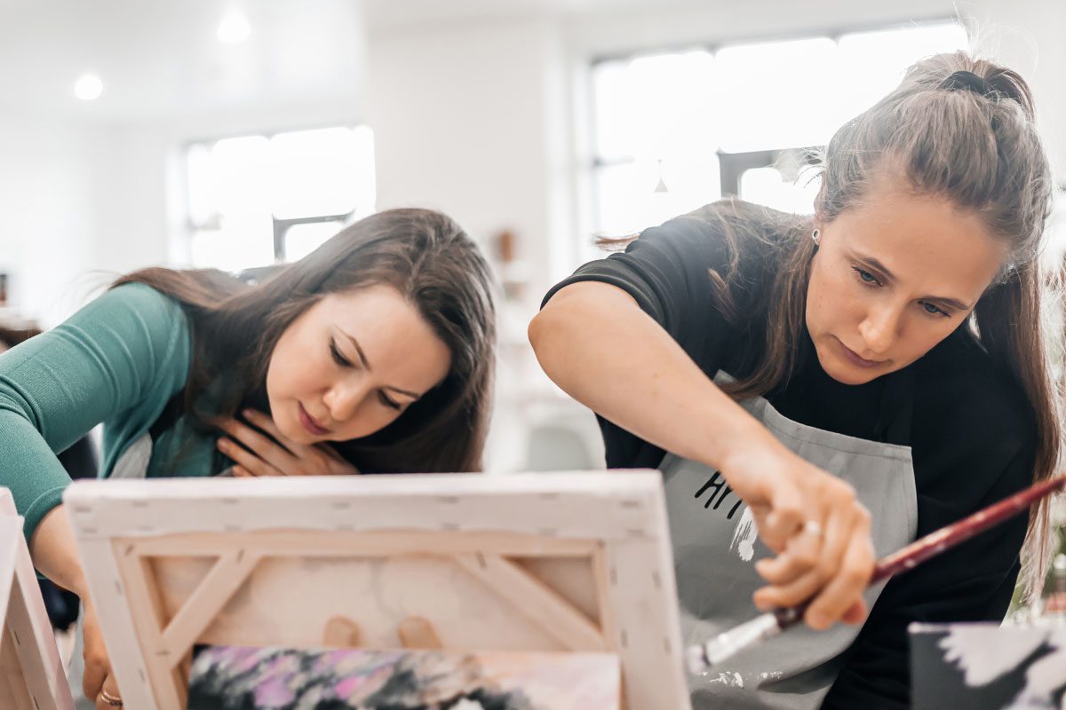 Two women are focused on painting at easels in the bright Greenway Lane Art Studio. One woman leans in closely, while the other applies paint with a brush, both appearing engaged in their artwork.