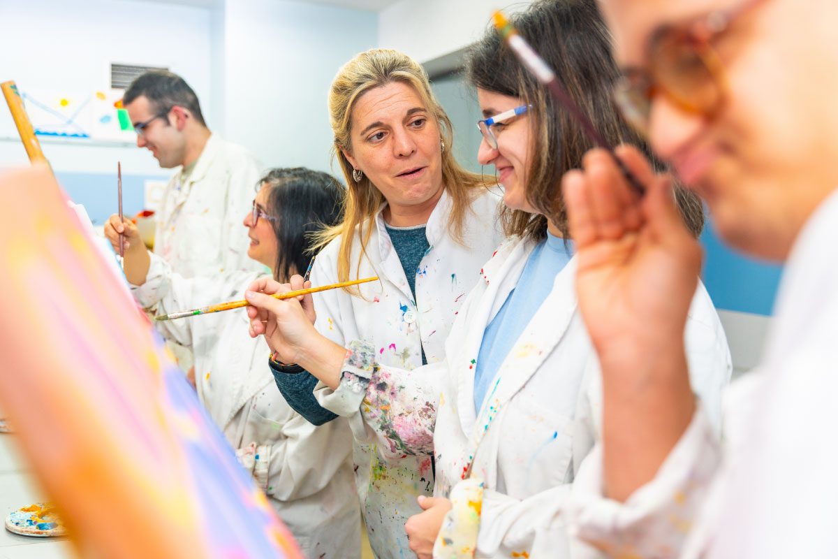 A group of adults wearing white smocks paint at easels in the bright Greenway Lane Art Studio. One woman smiles and speaks to another as they work, while others focus on their paintings.