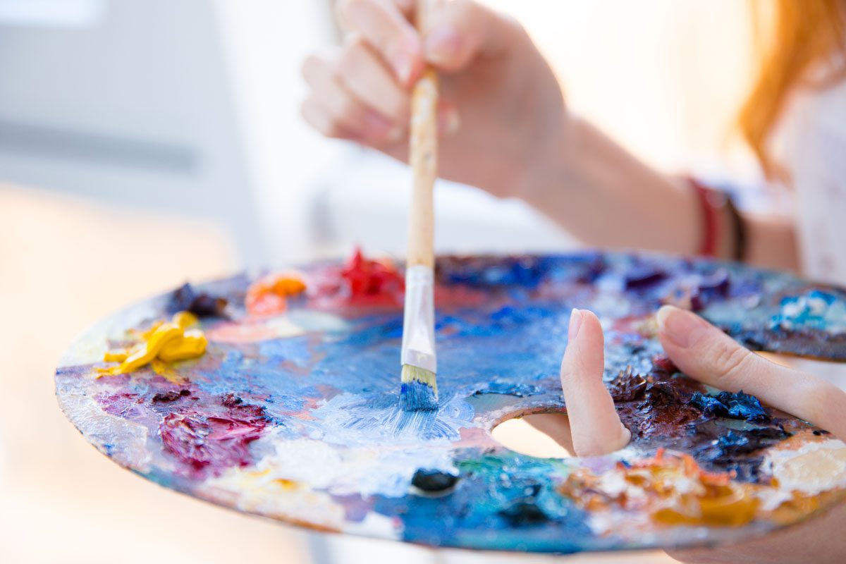 At Greenway Lane Art Studio, a person holds a colorful artist’s palette in one hand and uses a paintbrush with the other, mixing blue paint among various other vibrant colors.