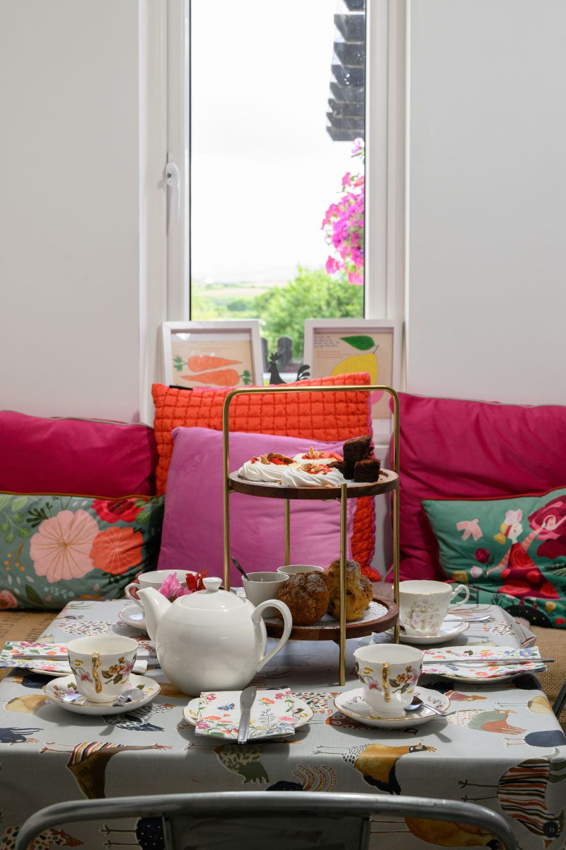 A cozy table set for tea at Greenway Lane Art Studio features a white teapot, floral teacups, plates, pastries, and cakes on a tiered stand. Colorful cushions surround the table, with a large window framing greenery and pink flowers outside.