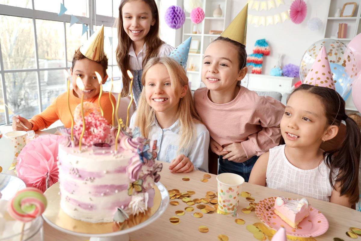 Five smiling children in party hats gather around a decorated birthday cake with candles at Greenway Lane Art Studio. Colorful decorations, balloons, and confetti fill the bright room, creating a festive atmosphere.