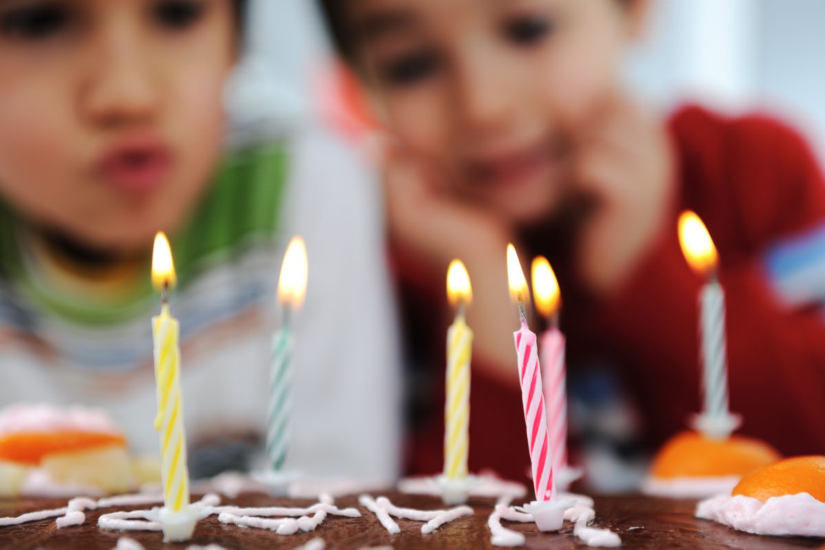 Two children eagerly watch a chocolate birthday cake decorated with five lit candles and orange slices at Greenway Lane Art Studio, ready to blow out the candles. The image is colorful and festive.