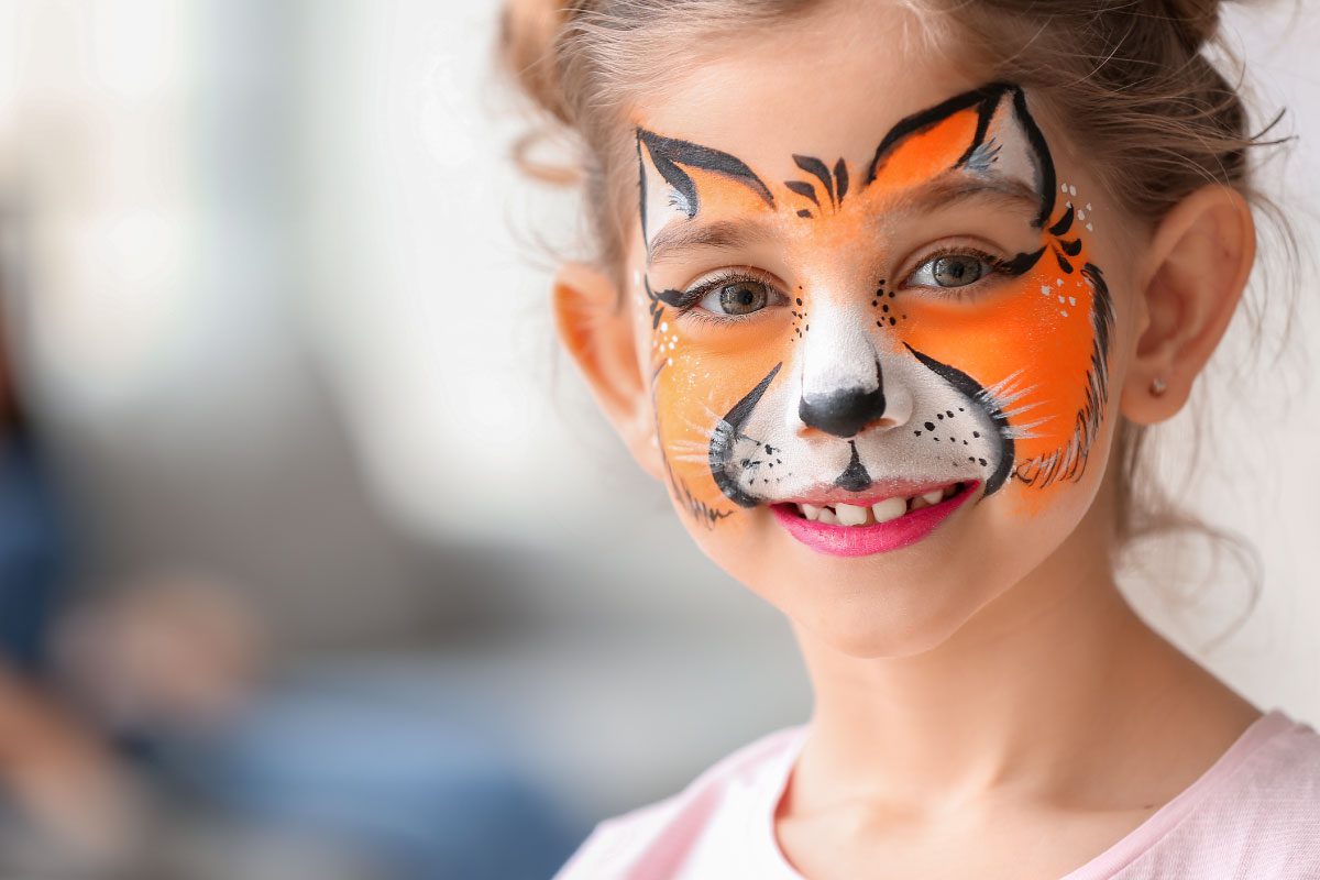 A young girl with light brown hair smiles, her face painted with a vibrant orange, black, and white tiger design at Greenway Lane Art Studio. She is indoors, wearing a light pink shirt, with a blurred background.