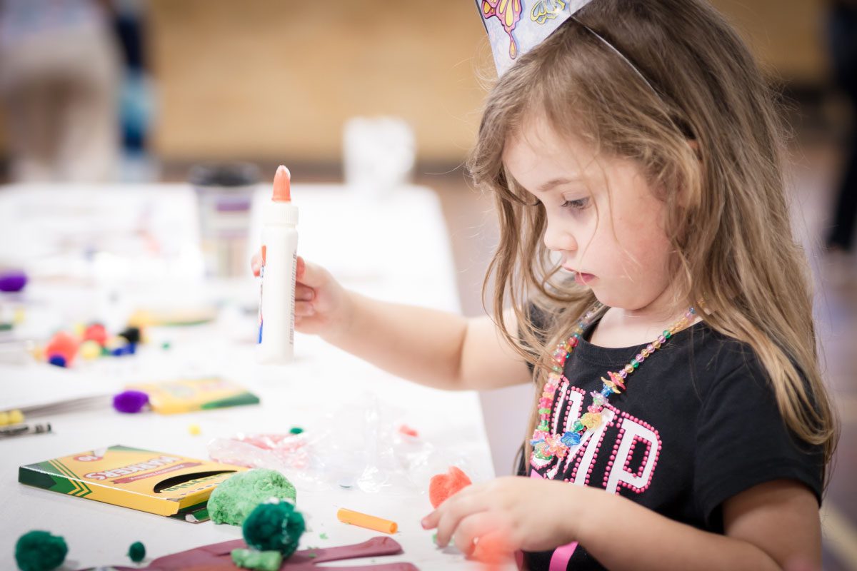 A young girl at Greenway Lane Art Studio, wearing a party hat and colorful necklace, concentrates on using a glue stick while doing arts and crafts at a table covered with crayons and pom-poms.
