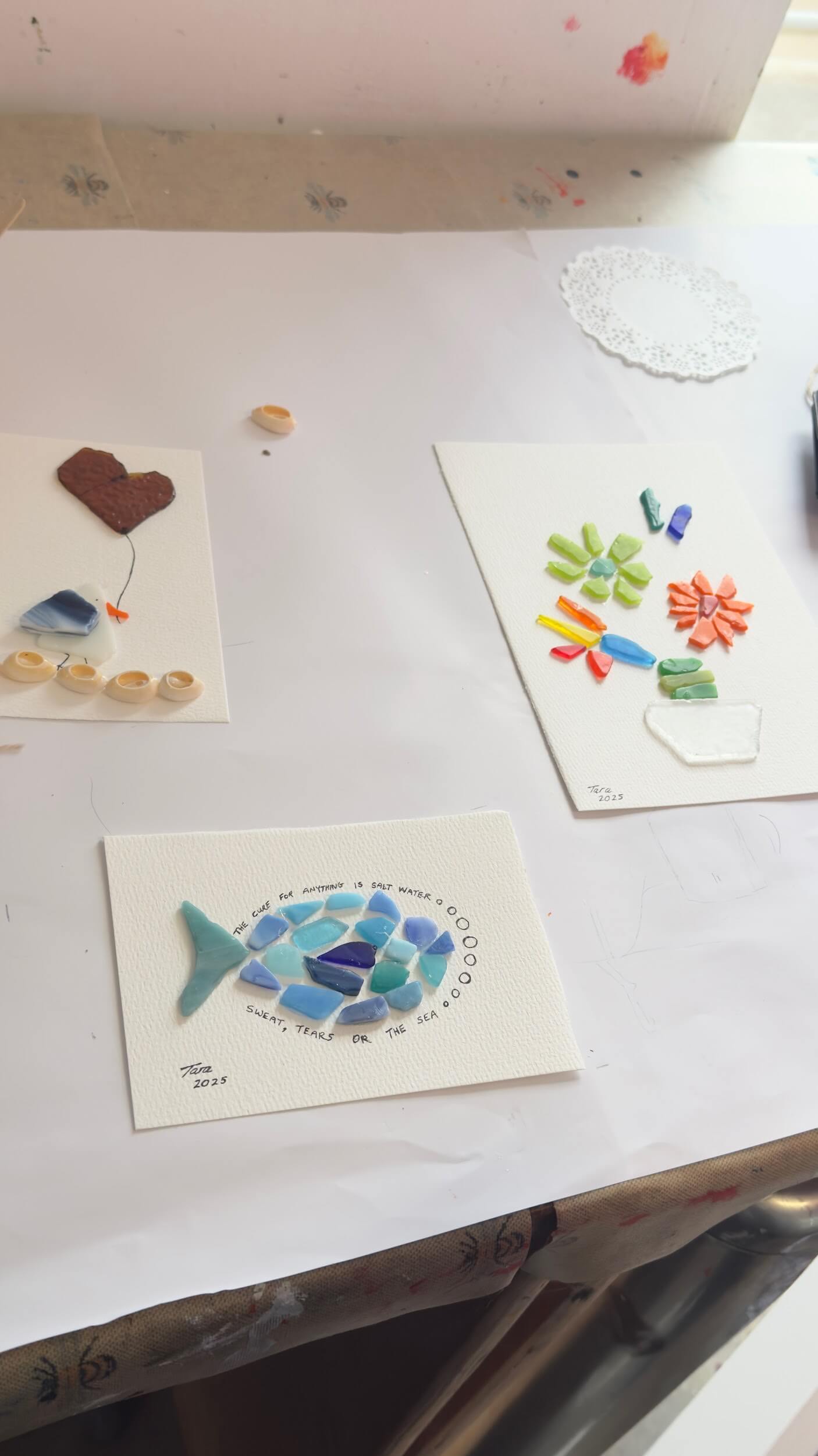 Three handmade art cards from Greenway Lane Art Studio sit on a table: one with a mosaic fish, one featuring a vase of colorful flowers, and another with a brown heart and blue shape—all crafted from sea glass and ceramic pieces.