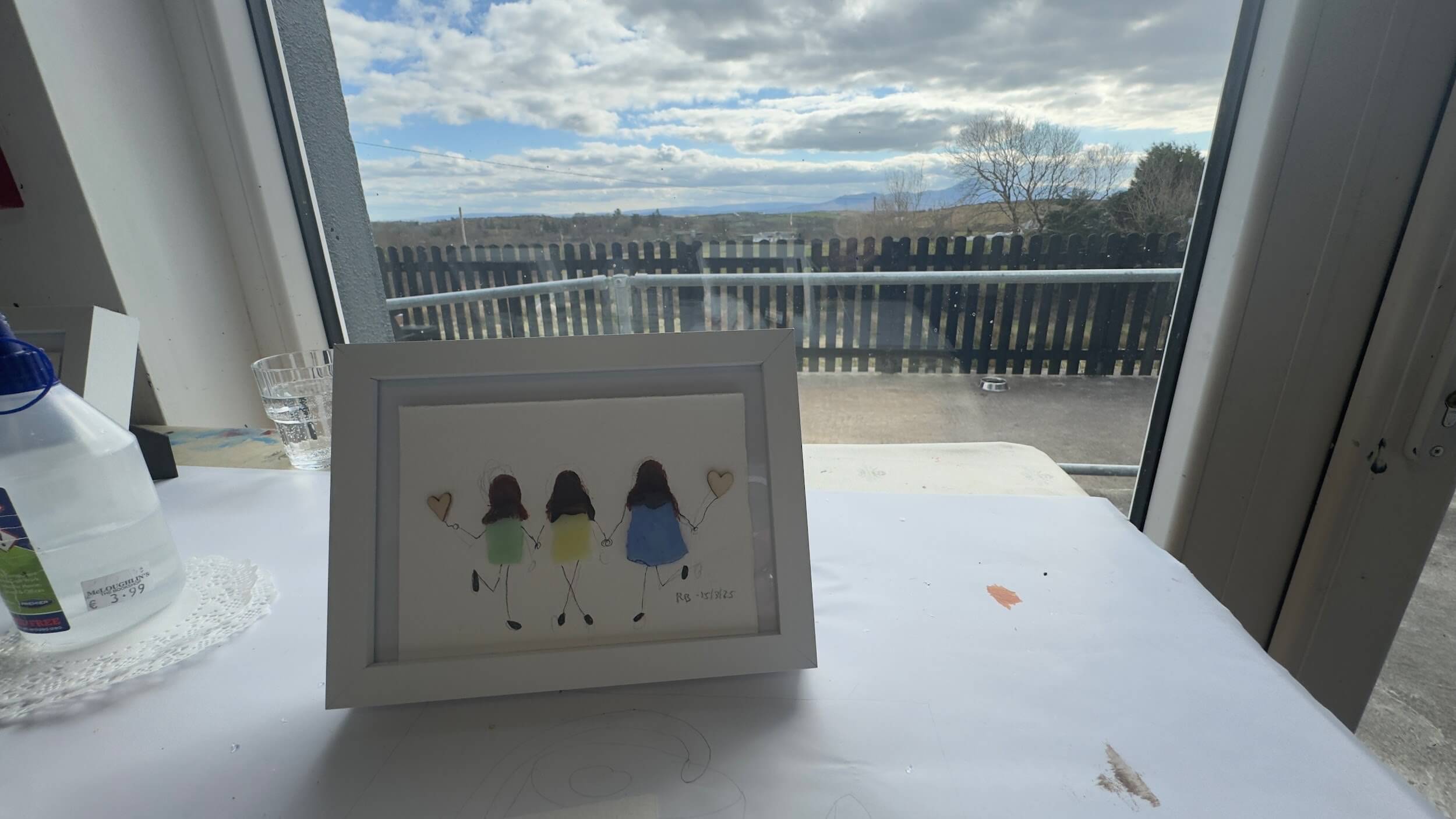 A framed drawing from Greenway Lane Art Studio depicts three girls holding heart-shaped balloons on a table by a large window, offering a scenic view of trees, mountains, and a cloudy sky. Nearby sit a water bottle and glass.