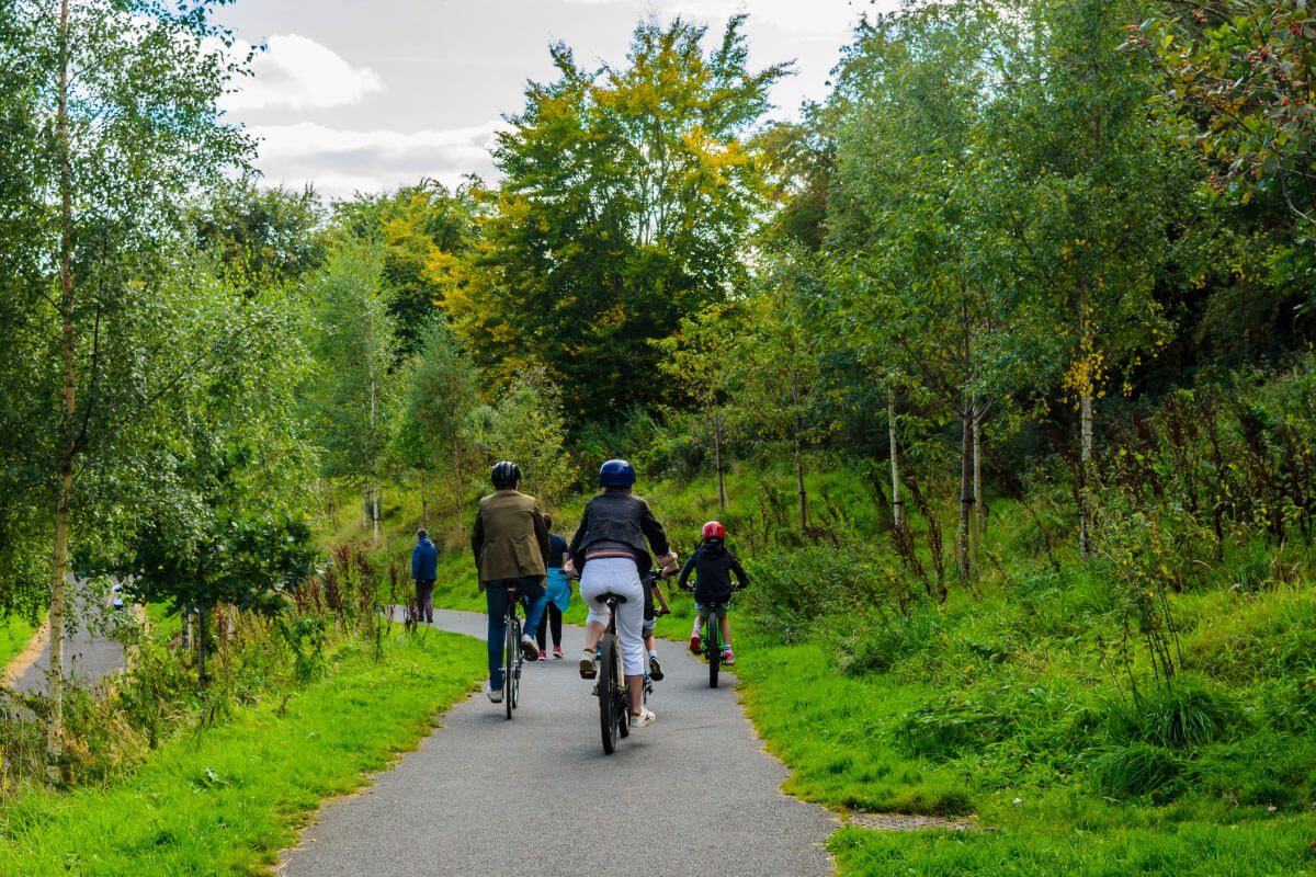 A group of people, including adults and children wearing helmets, ride bicycles on a paved path through a lush green park near Greenway Lane Art Studio on a partly cloudy day.