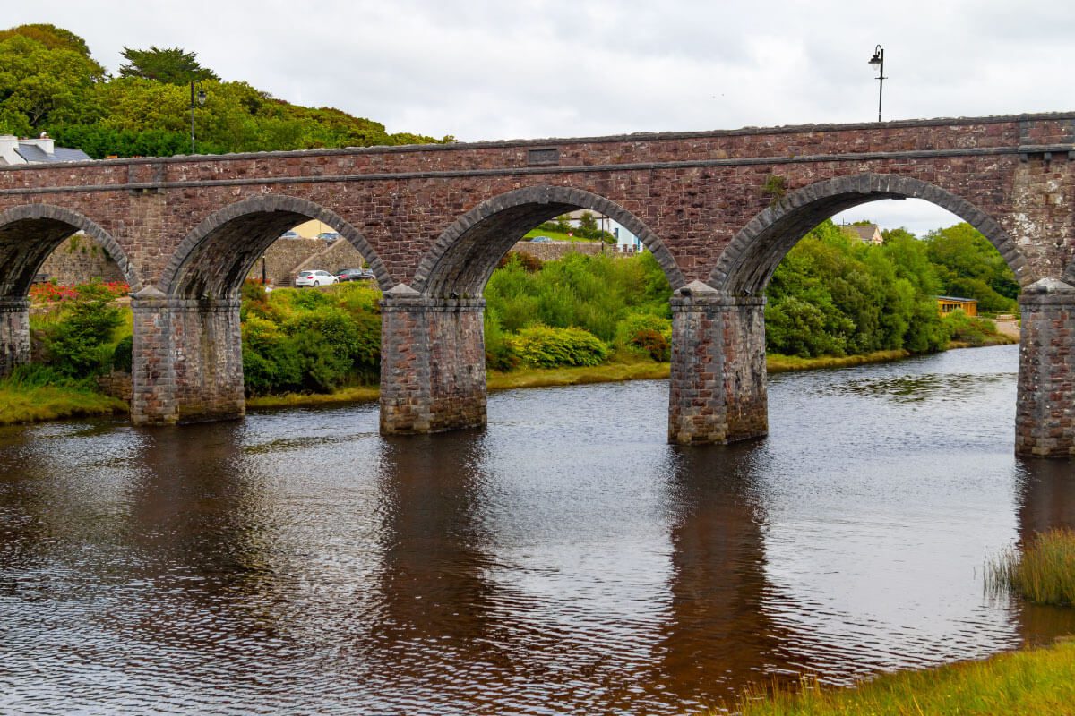 A stone arch bridge with four arches spans over a calm river, surrounded by green shrubs and trees near Greenway Lane Art Studio, under a cloudy sky.
