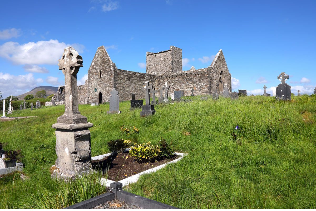Ancient stone church ruins on Greenway Lane Art Studio grounds, surrounded by a grassy cemetery with several Celtic cross gravestones under a bright blue sky with scattered clouds.