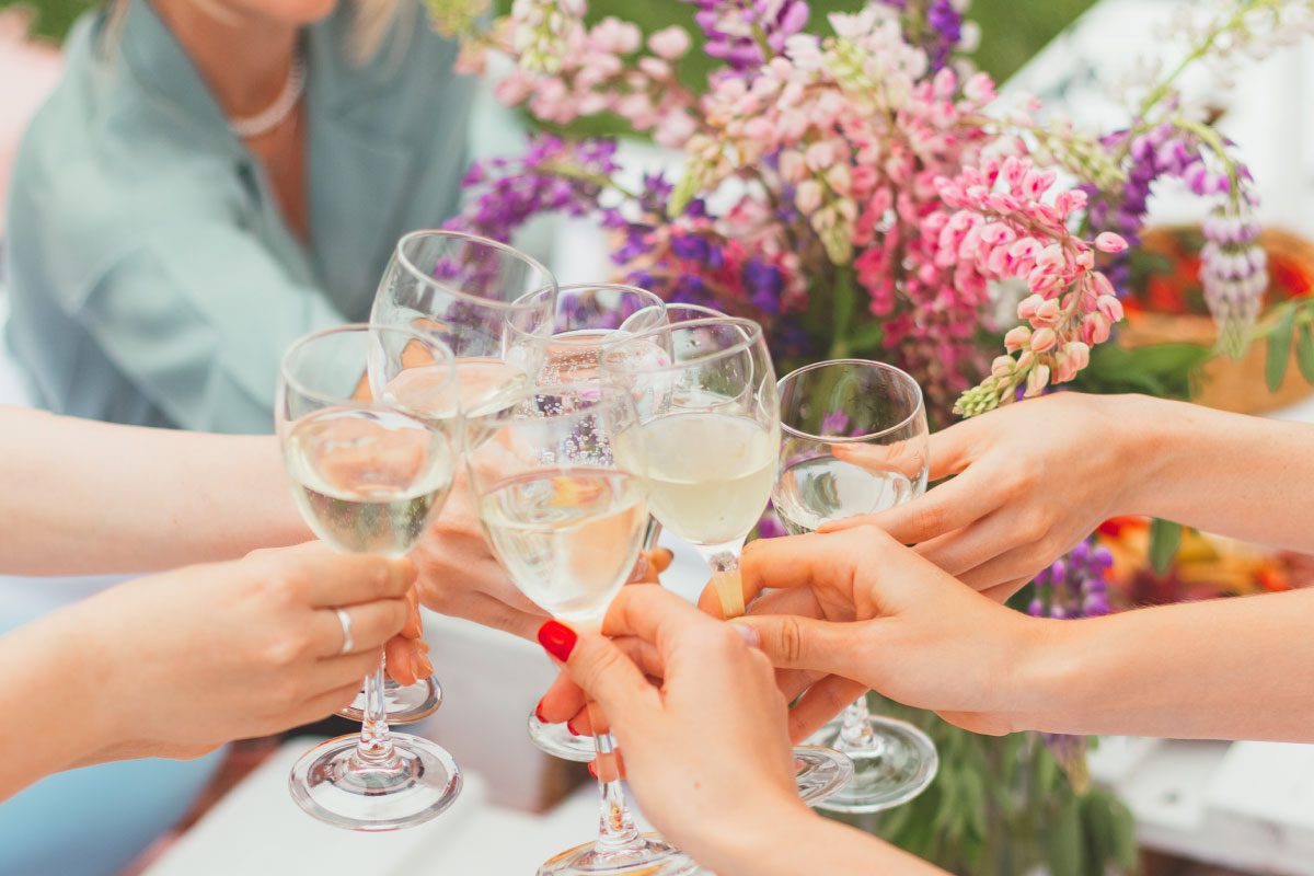 A group of people clinking glasses of white wine in a toast, surrounded by colorful flowers on a sunny outdoor table at Greenway Lane Art Studio. The focus is on the hands and glasses, with one person blurred in the background.