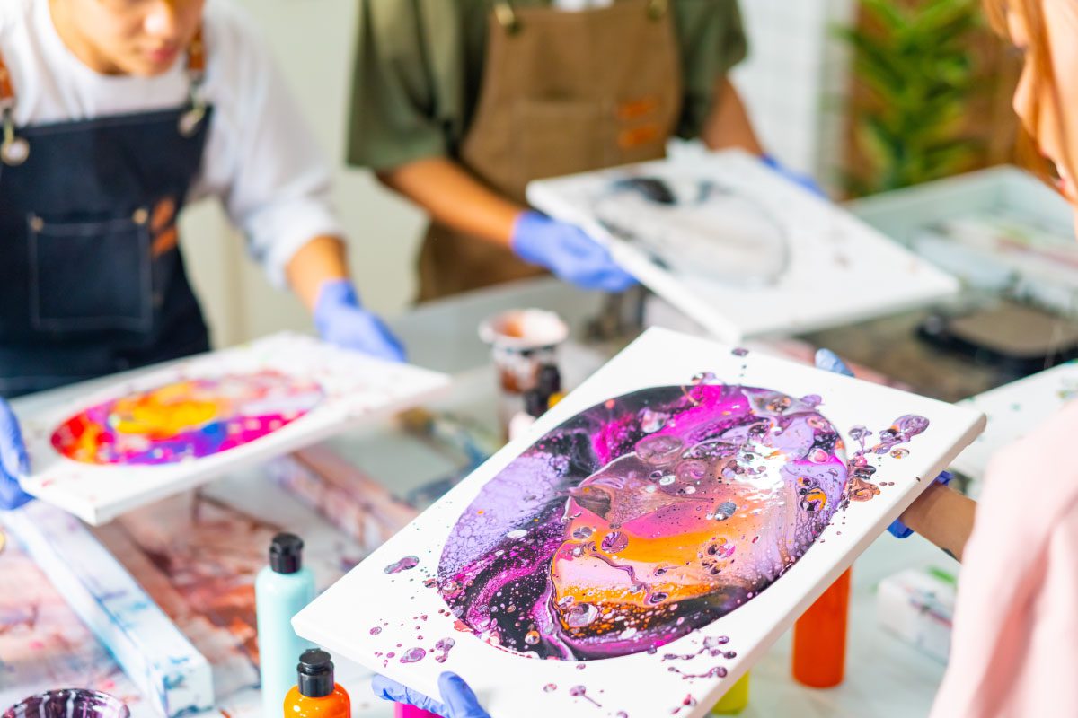 At Greenway Lane Art Studio, three people in aprons and gloves proudly hold abstract acrylic pour paintings bursting with vibrant purple, pink, and orange hues at a table filled with art supplies and paint bottles.