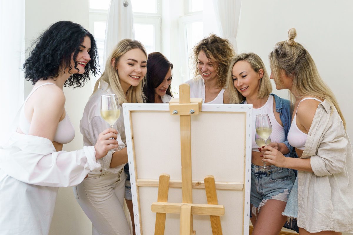 Six young women stand together smiling and holding glasses of white wine at Greenway Lane Art Studio, gathered around a canvas on an easel in a bright room, enjoying a fun painting activity or art class.