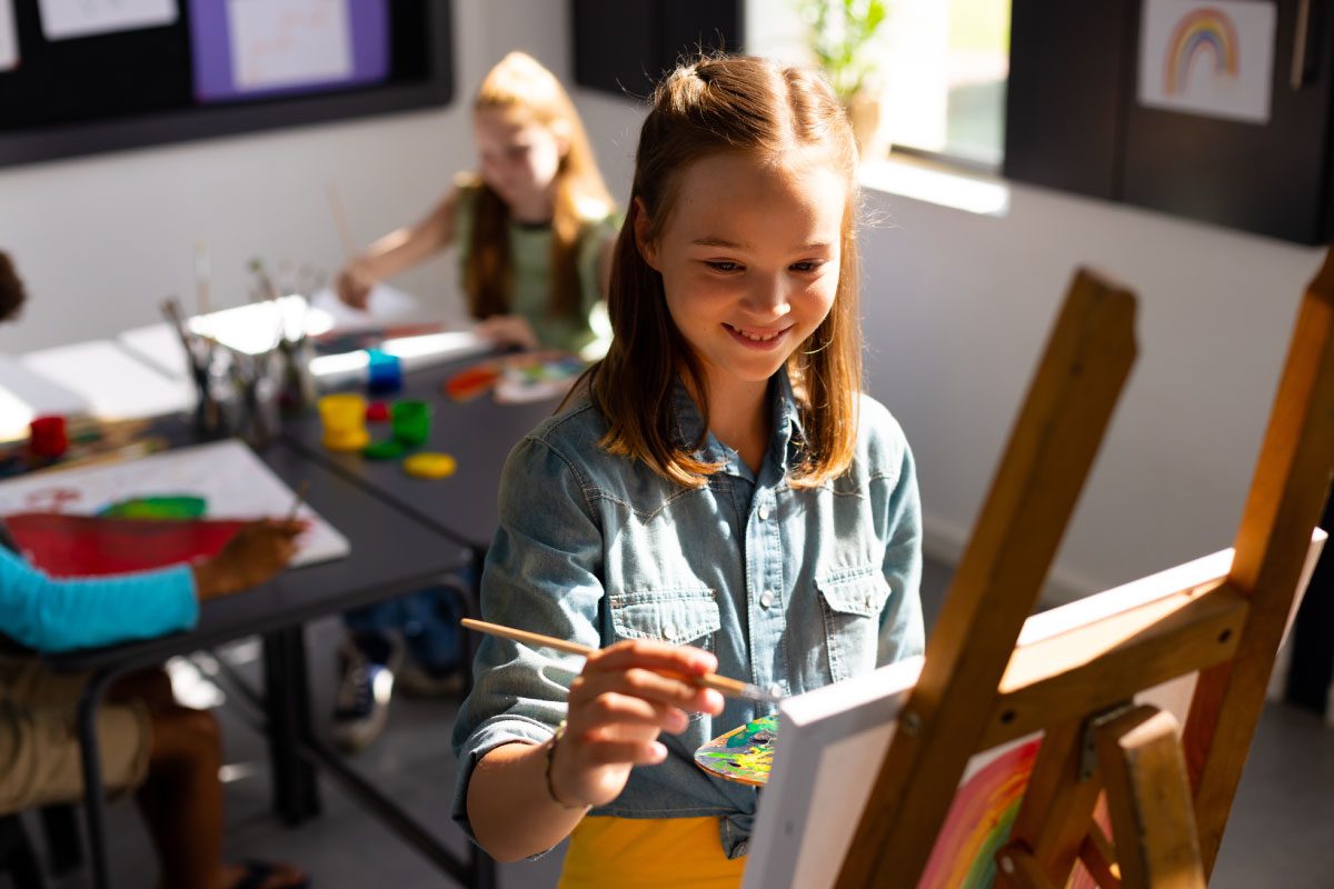 A smiling girl paints on a canvas at an easel in a bright Greenway Lane Art Studio classroom, while other children create art at a table in the background. Art supplies and colorful paintings are visible around the room.