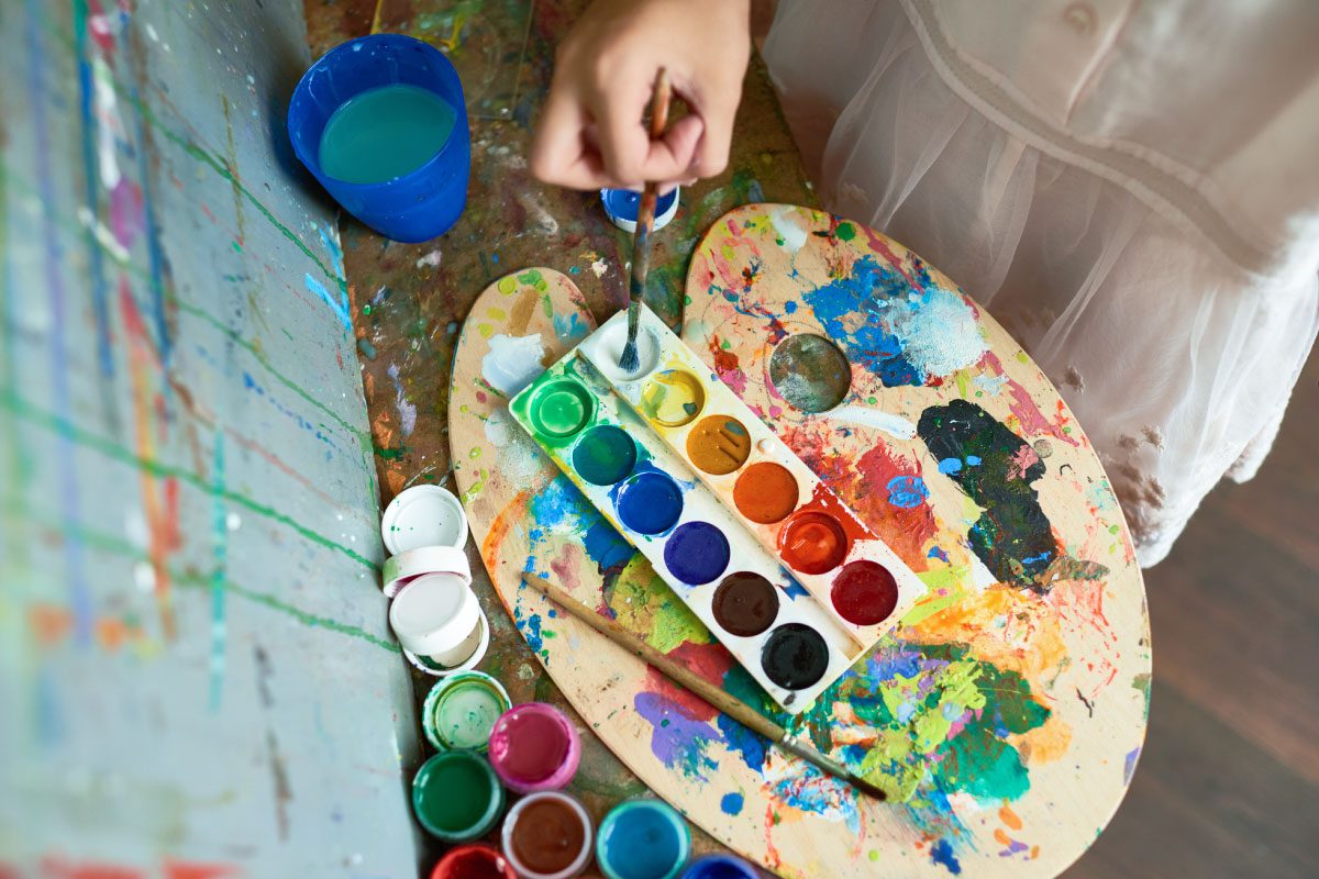 At Greenway Lane Art Studio, a person in a white dress dips a paintbrush into a water cup beside a colorful watercolor palette and paint-splattered artist’s palette, with jars of paint on a messy art table.
