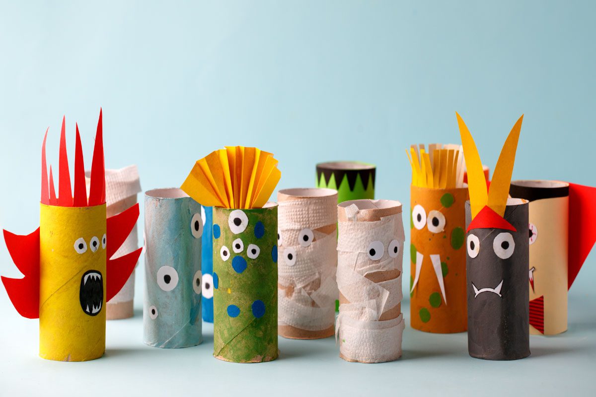 A group of colorful toilet paper rolls decorated as quirky monsters with googly eyes, painted designs, and paper accents like spikes, horns, and fins, arranged on a light blue background at Greenway Lane Art Studio.