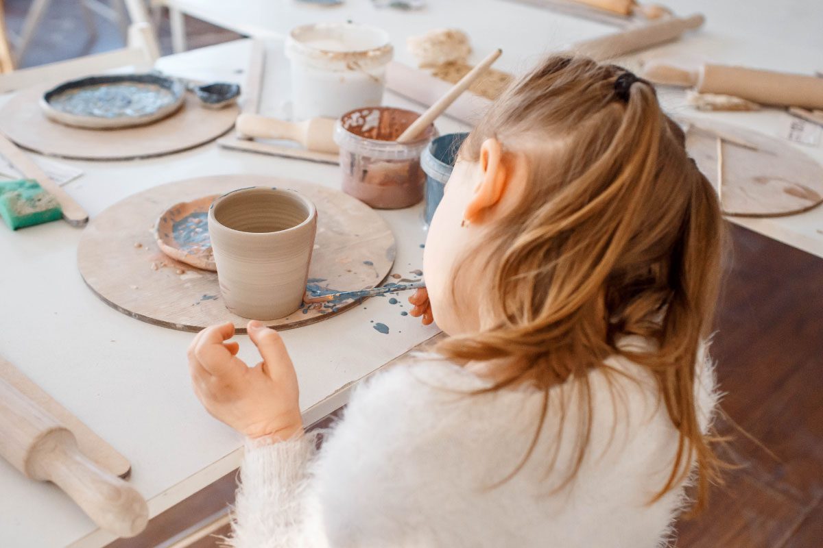 A young girl with light brown hair sits at a table, painting a clay cup during a pottery class at Greenway Lane Art Studio. Art supplies, paint, and clay tools are scattered on the table around her.
