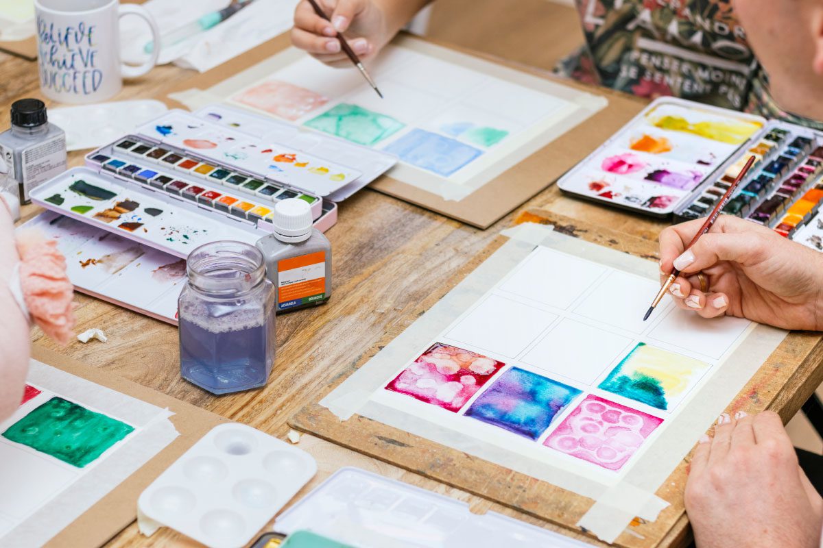Three people sit around a table at Greenway Lane Art Studio, painting colorful watercolor swatches on paper. Art supplies, including palettes, brushes, and jars of water, are spread across the wooden surface.