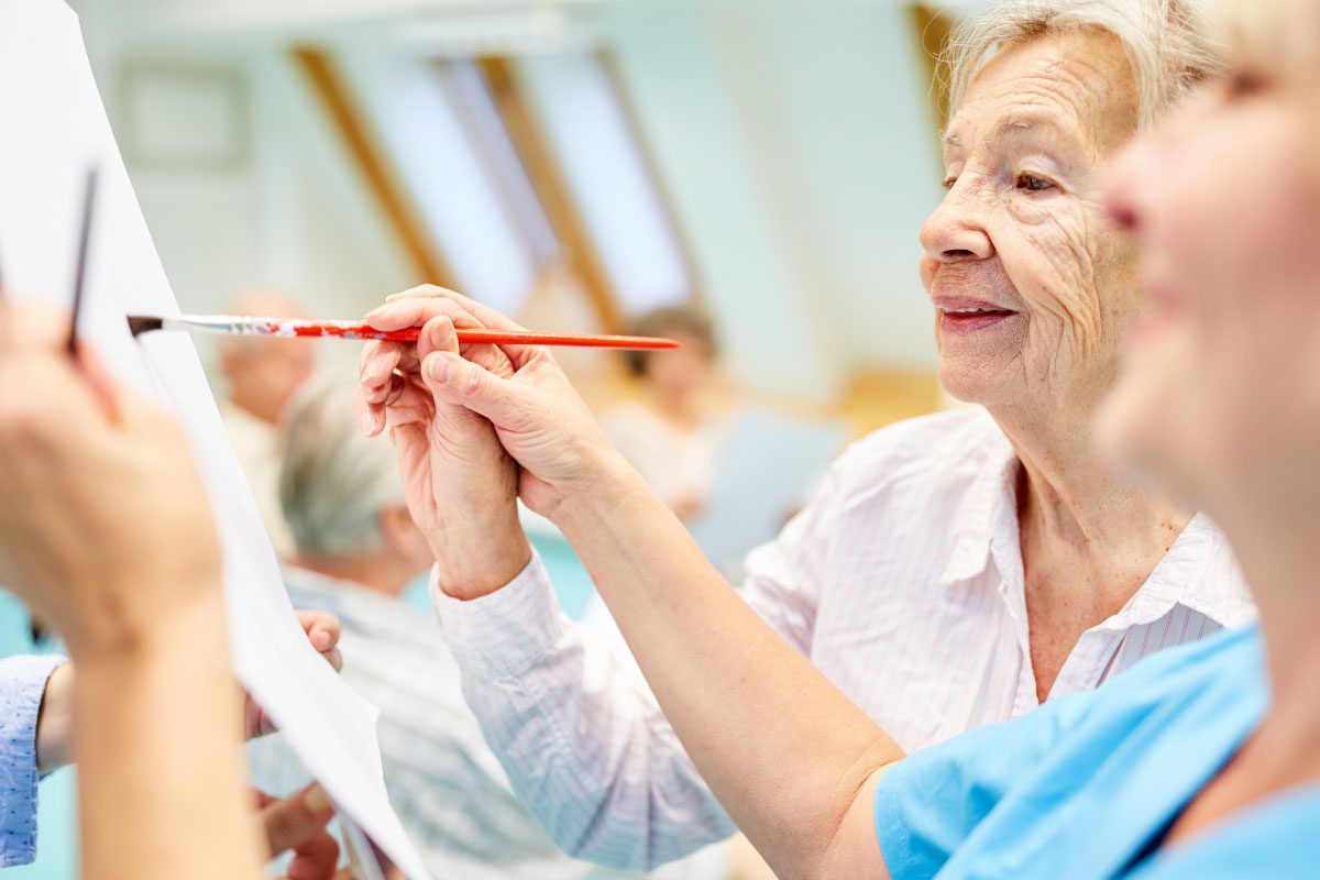 An elderly woman smiles as she paints on a canvas with assistance from a caregiver at Greenway Lane Art Studio. Both are holding paintbrushes, suggesting a shared art activity in a bright, cheerful room.