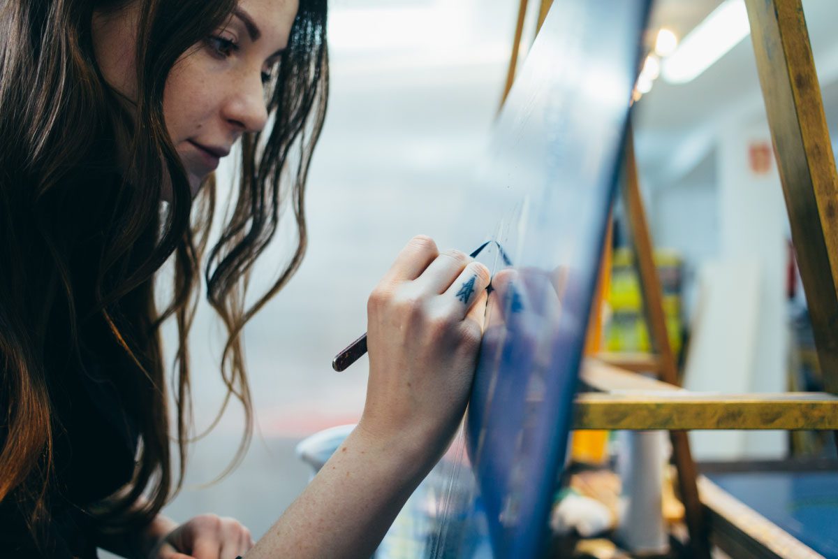 A woman with long brown hair is focused on painting a canvas set on an easel at Greenway Lane Art Studio. She holds a paintbrush and works on a detailed section of the artwork in a brightly lit room.