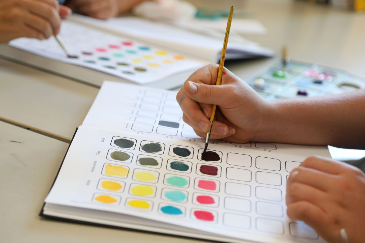 At Greenway Lane Art Studio, a person uses a paintbrush to fill a grid with colors in a sketchbook, likely creating a color chart. Another hand paints in the background, with watercolor paints and palettes on the table.