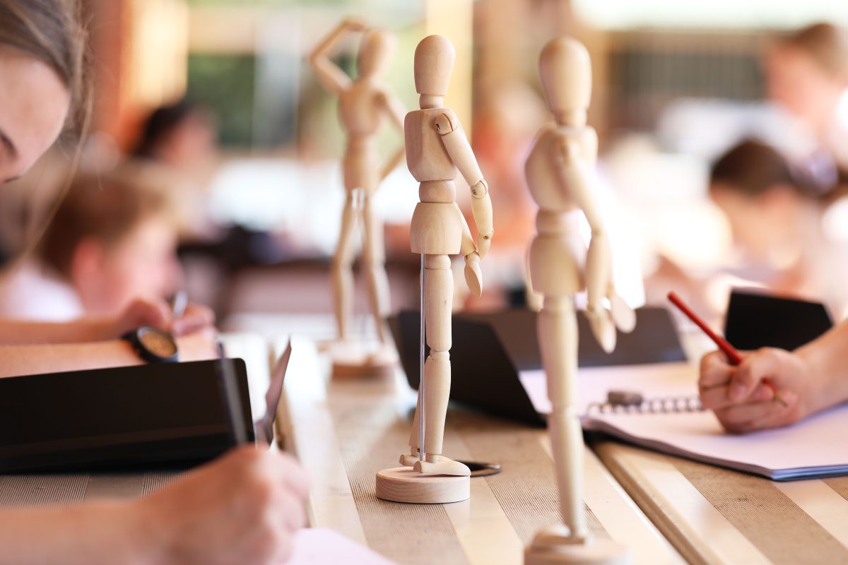 Wooden artist mannequins are placed on a table among sketchbooks and people drawing, suggesting an art class setting at Greenway Lane Art Studio with students focused on sketching.