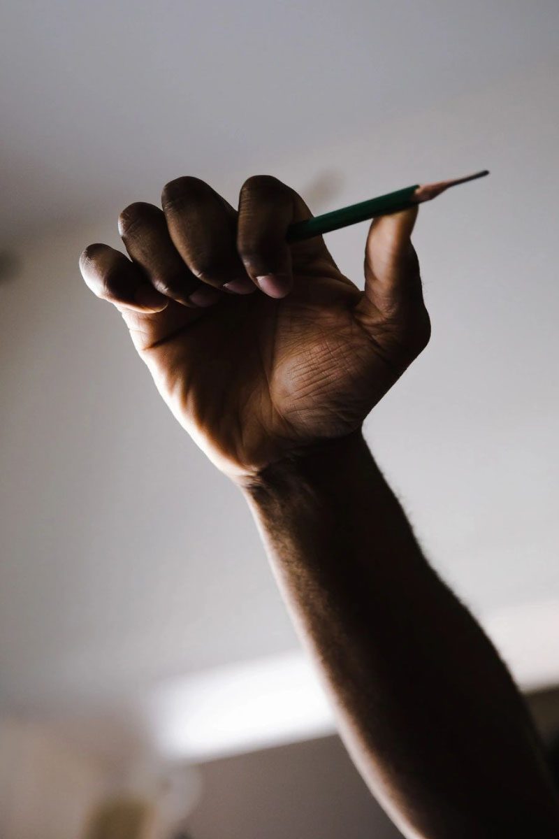 A raised hand holds a green pencil against a blurred, light background, suggesting creativity, power, or expression—a perfect capture of the inspiration found at Greenway Lane Art Studio. The low angle emphasizes the hand and pencil.