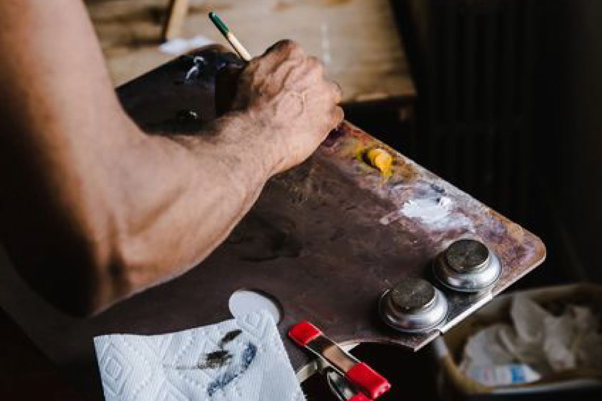 A person’s hand holding a paintbrush, working on a wooden palette with paint dabs and metal containers at Greenway Lane Art Studio, with a paper towel and red clamp attached to the edge.