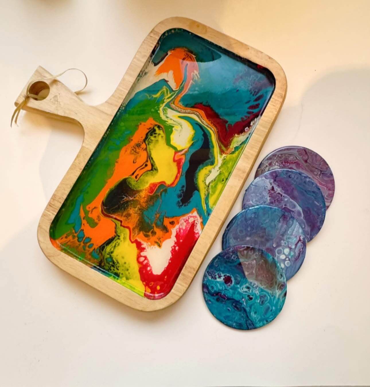 A wooden serving tray with a colorful, abstract resin design by Greenway Lane Art Studio is placed beside three round coasters with blue and purple swirl patterns on a white surface.