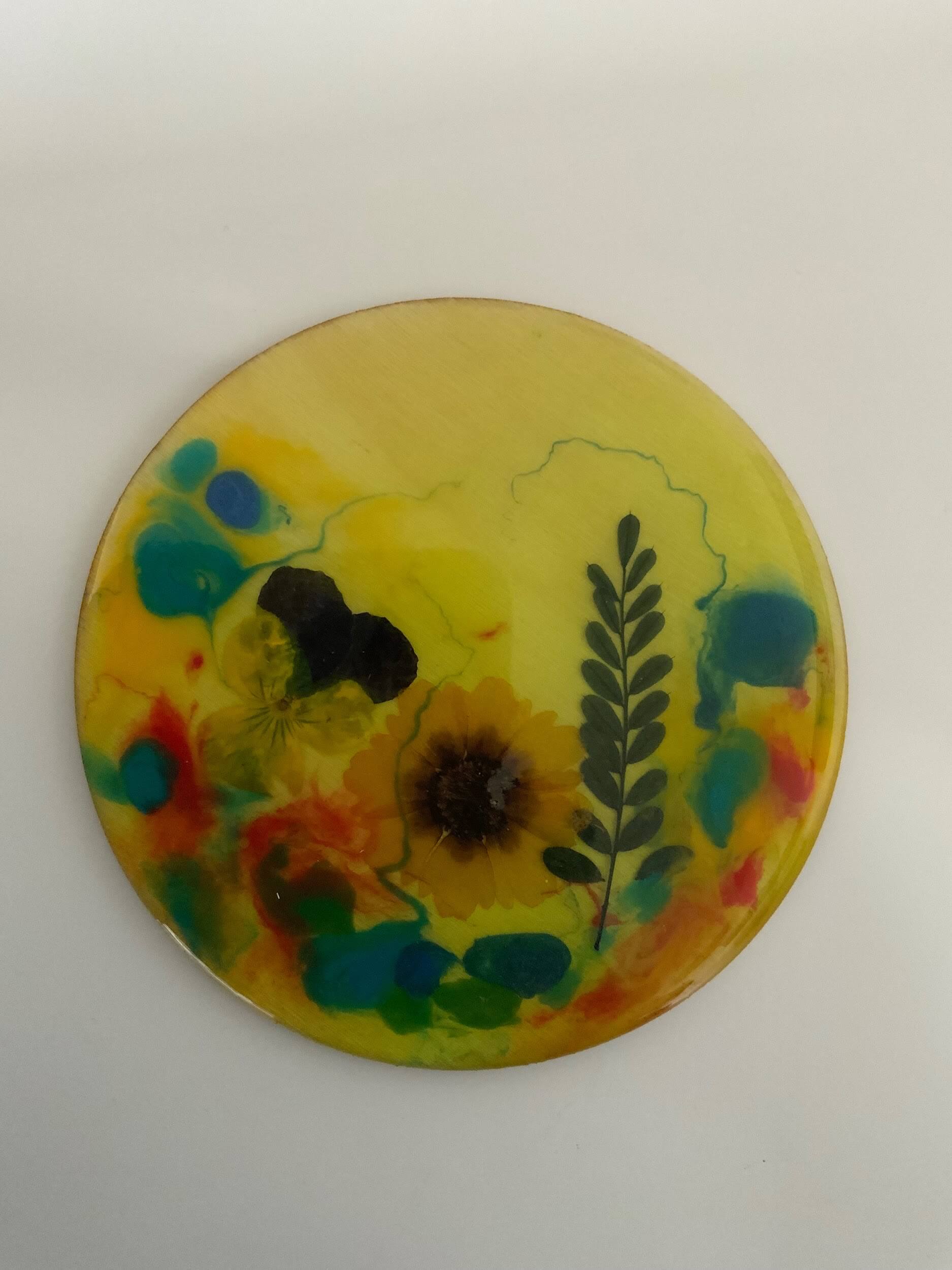 A round coaster with a yellow background, decorated with a dried flower, a fern leaf, and colorful abstract spots in blue, green, red, and orange—crafted by Greenway Lane Art Studio.
