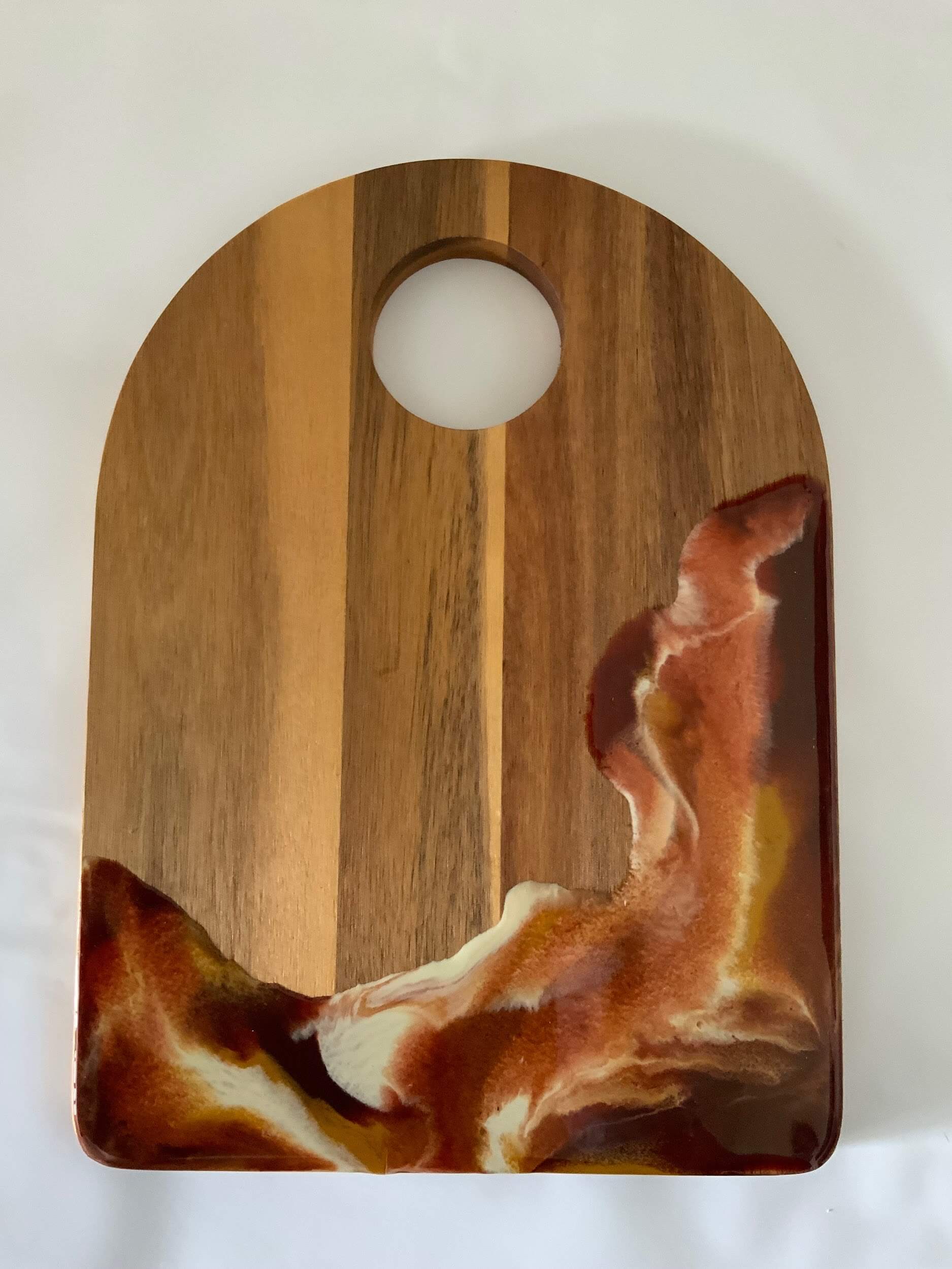 A wooden cutting board with a rounded top and a circular hole near the top. The lower right corner features a decorative resin design in swirling shades of brown, white, and rust by Greenway Lane Art Studio.