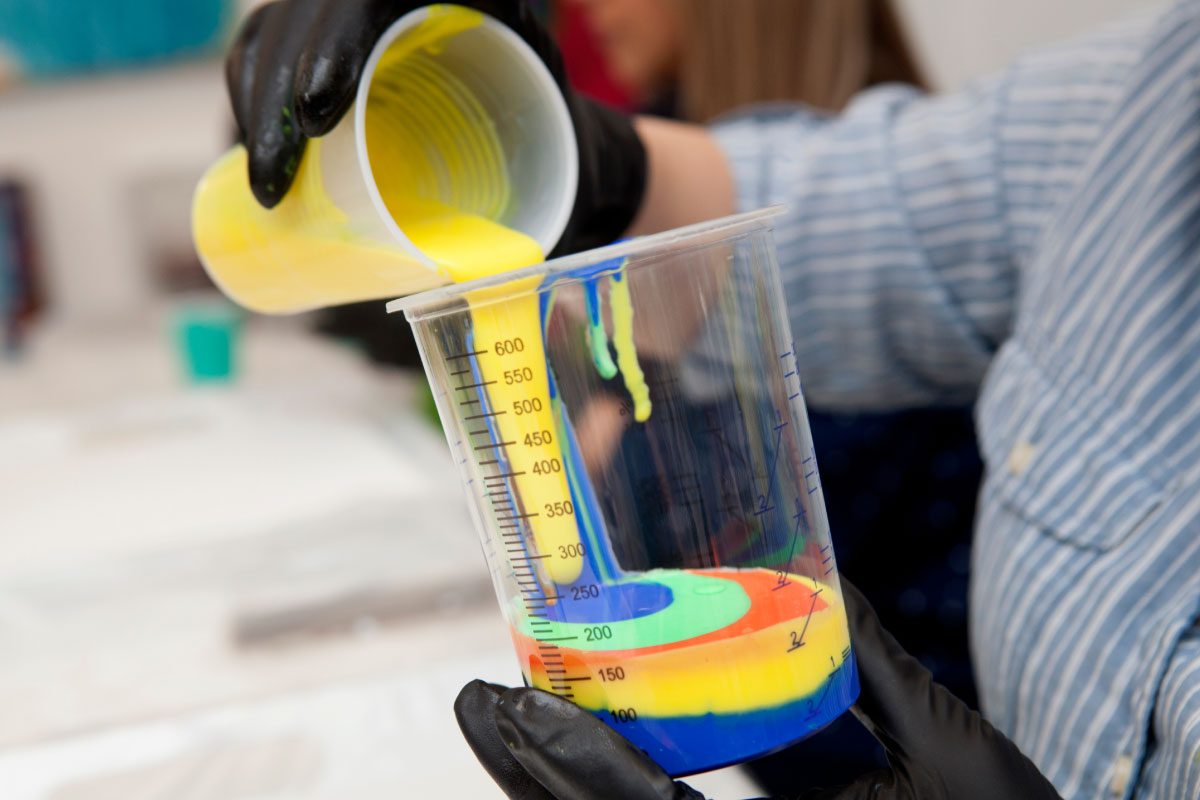 At Greenway Lane Art Studio, a person wearing black gloves pours yellow paint into a cup with blue, green, and red paints, creating colorful layers. The clear plastic cups show off the vibrant mixes with measurement markings.