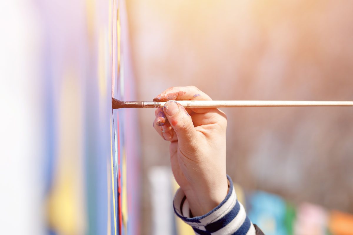 A close-up of a hand holding a paintbrush, painting on a vertical surface outdoors. The hand is covered in paint, and the blurred background hints at Greenway Lane Art Studio bathed in soft sunlight and indistinct shapes.