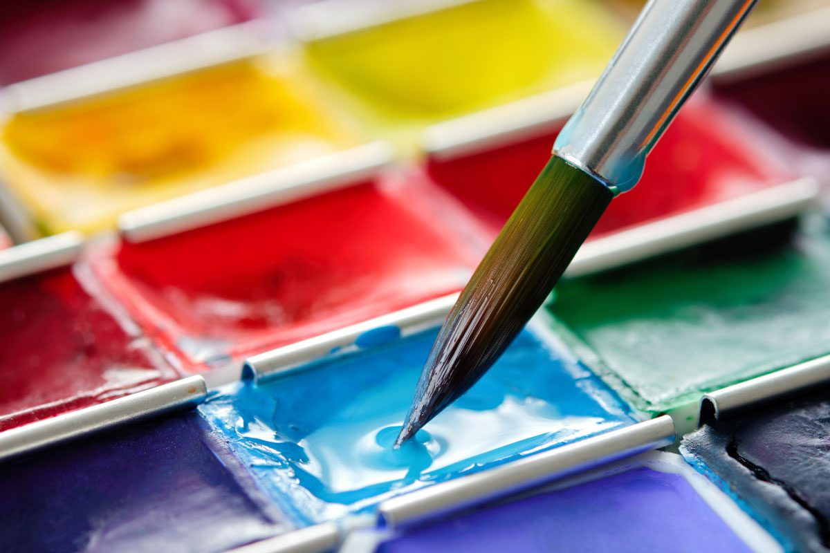 A close-up of a paintbrush touching a blue watercolor pan in a palette at Greenway Lane Art Studio, surrounded by various vibrant colors such as red, yellow, and green.