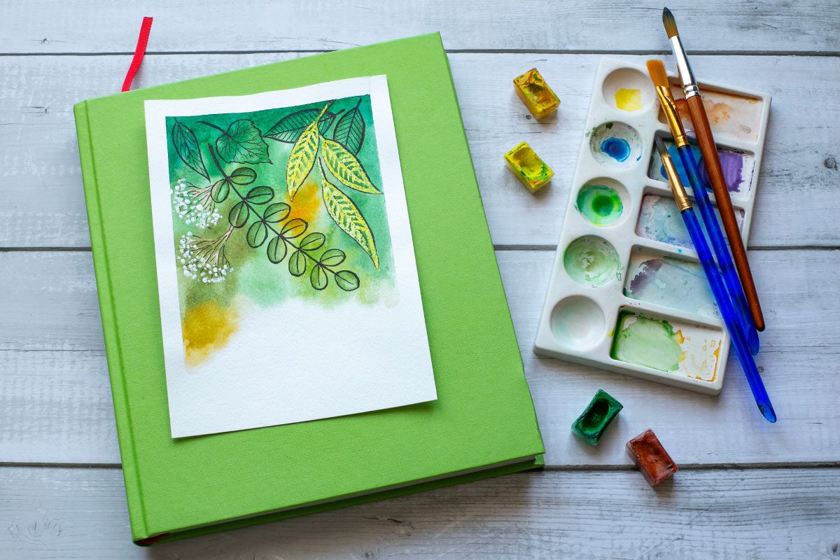 Watercolor painting of leaves on white paper rests on a green book next to a palette with paint, brushes, and watercolor pans on a white wooden surface at Greenway Lane Art Studio.